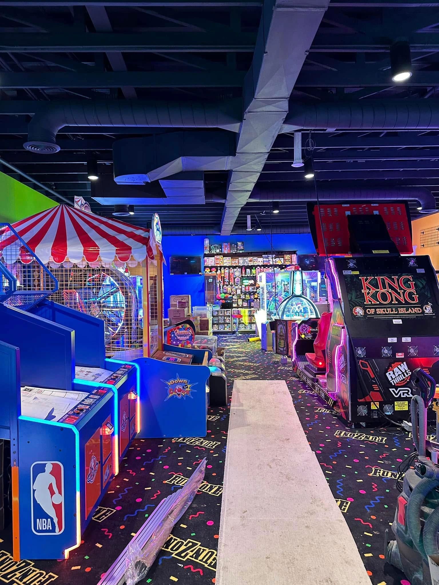 Arcade with rows of game machines, a midway game, and a colorful patterned floor. Dim lighting.
