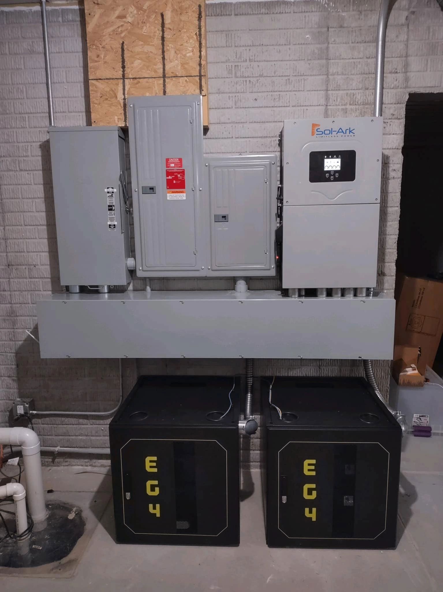 Electrical components and battery storage units mounted on a basement wall. Grey panels and two black EG4 batteries.