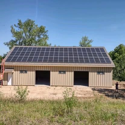 Solar panels on a tan barn with two open doors. A person stands on the right. Blue sky.