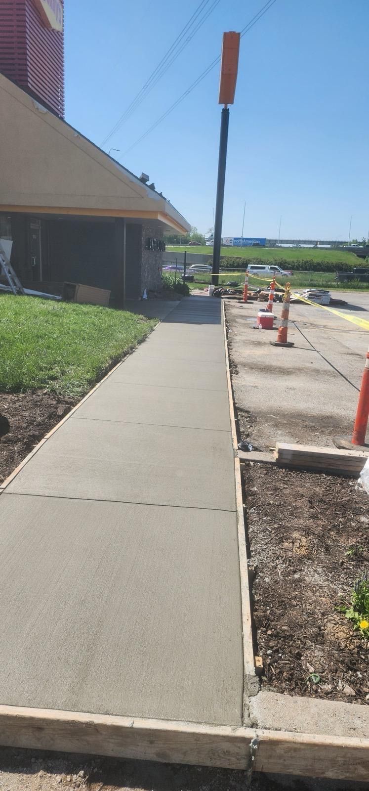 Concrete sidewalk beside a building, leading past a signpost and orange cones in a construction area