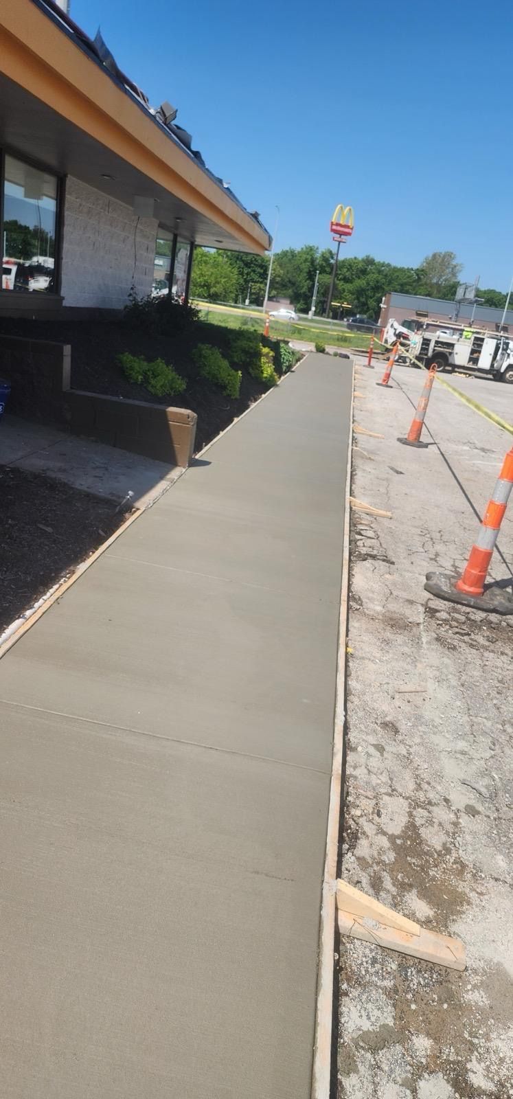 Sidewalk beside a storefront under a blue sky, with orange cones lining a gravel construction area.