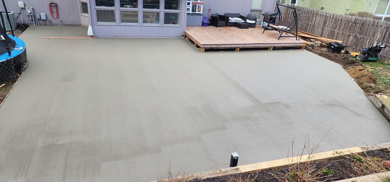 Freshly poured concrete patio beside a house with a small wooden deck and glass doors.