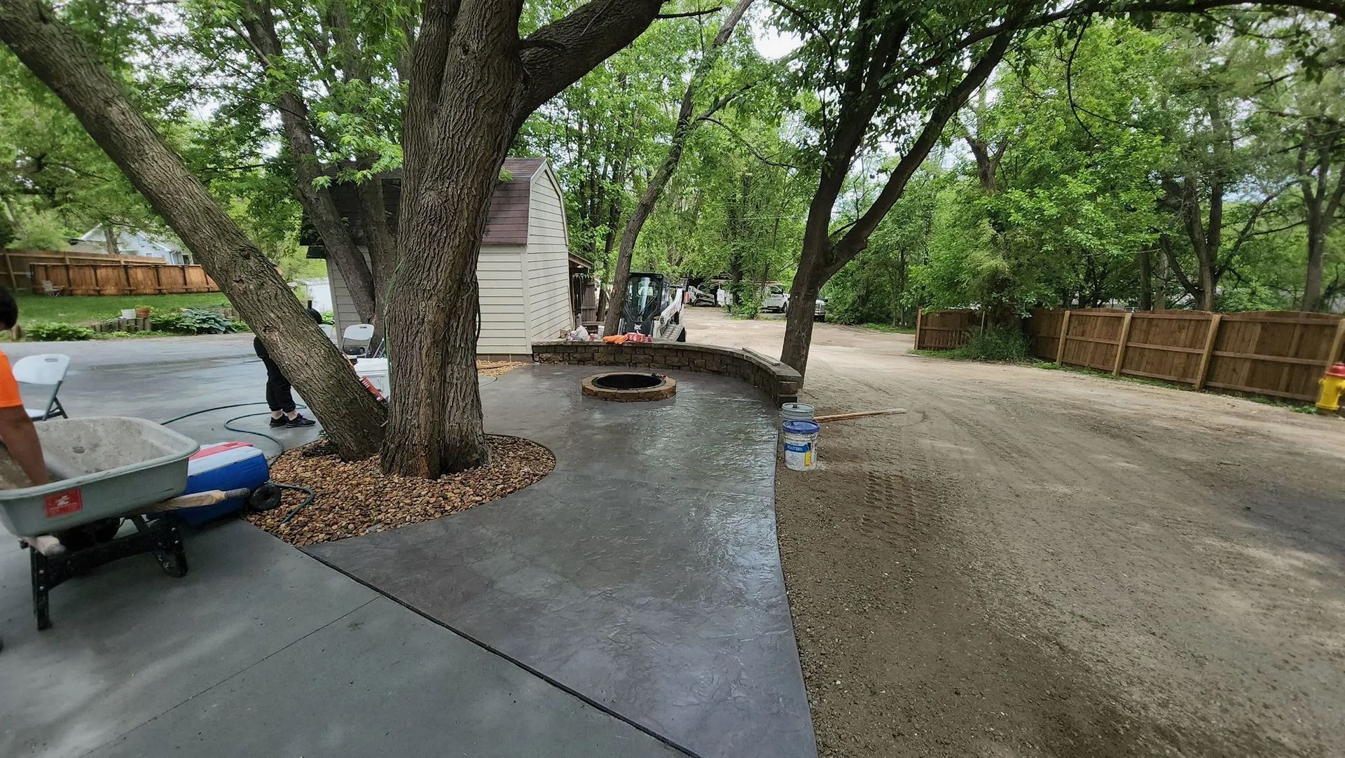 Tree-shaded backyard patio with curved concrete path, fire pit, and gravel driveway
