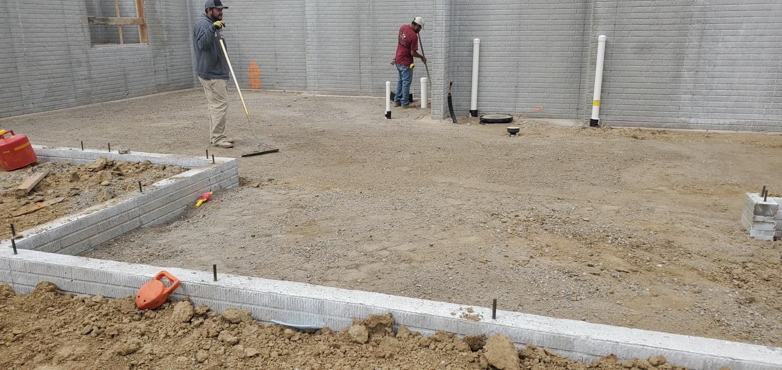 Construction site with workers leveling gravel beside concrete foundations and a retaining wall