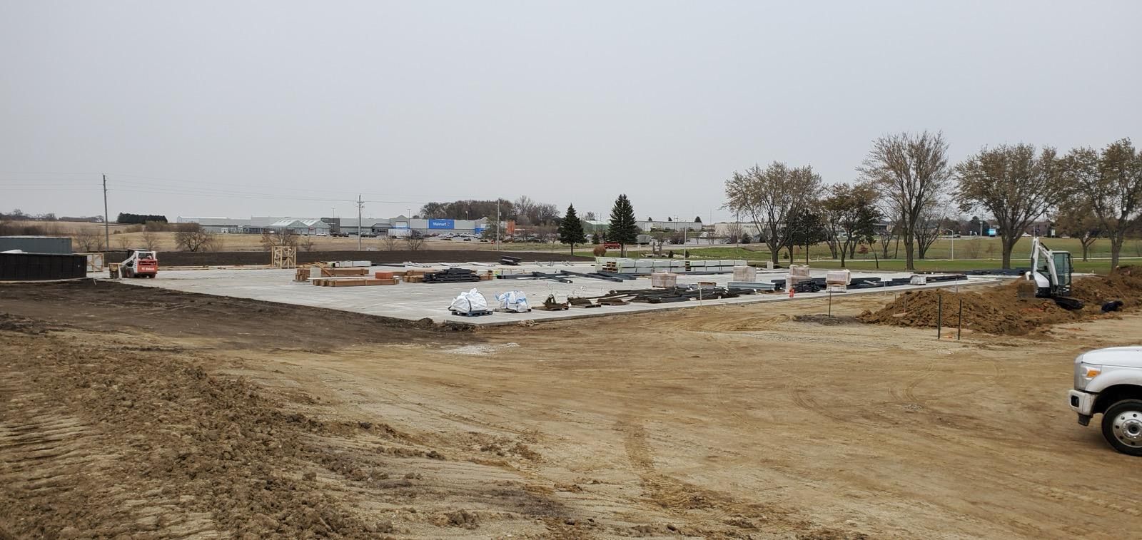 Wide muddy construction site with concrete foundations, white materials, and trees in the background