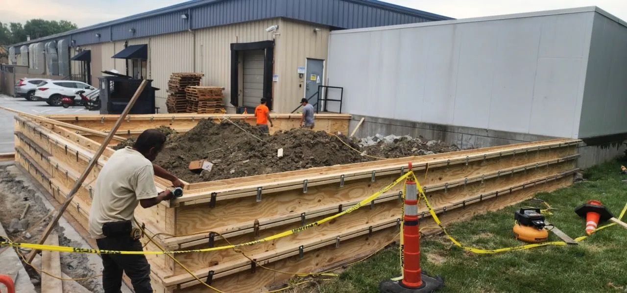 Construction crew installing wooden retaining wall beside a building, with orange cones and excavator tracks visible.