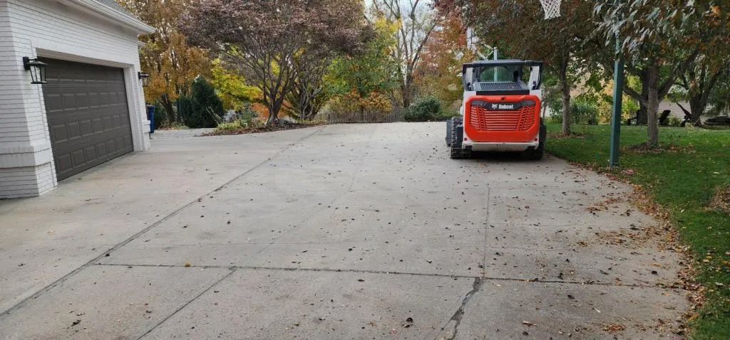 Orange skid steer parked on a concrete driveway beside a garage and trees