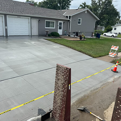 Fresh concrete driveway beside a gray house, with caution tape, orange cones, and work tools in front.