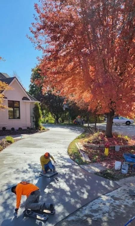 Two workers paving a curved driveway beside a house and autumn trees