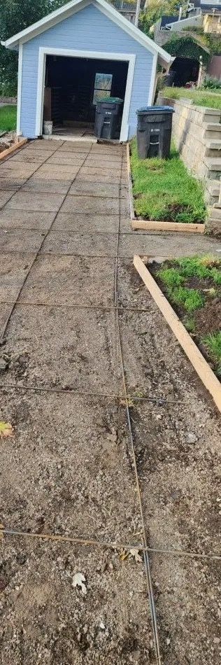 Concrete walkway leading to a blue garage with open door, bordered by garden beds and a trash bin