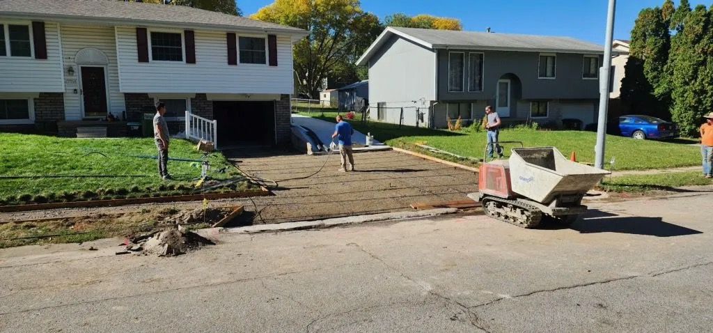 Two workers shoveling dirt by a sidewalk in front of houses, with a wheelbarrow on the street.