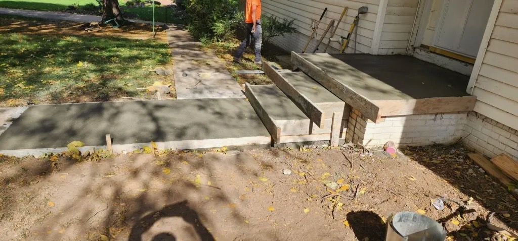 Concrete walkway with steps beside a house, bordered by a small yard and trees.