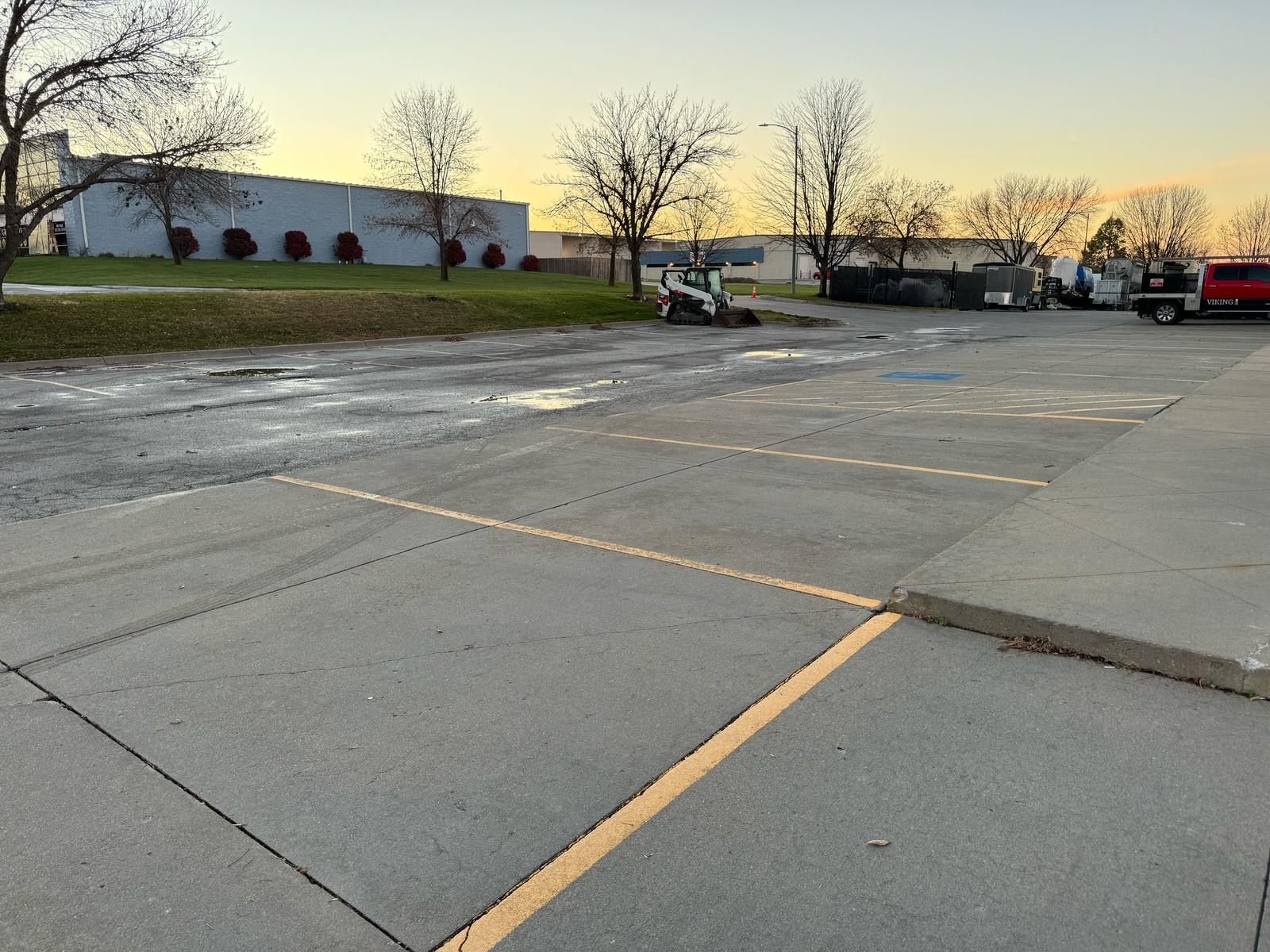 Empty parking lot at sunset with cracked pavement, leafless trees, and industrial buildings in the distance
