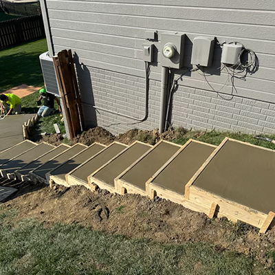 Concrete steps under construction beside a gray house, with soil and utility boxes nearby.