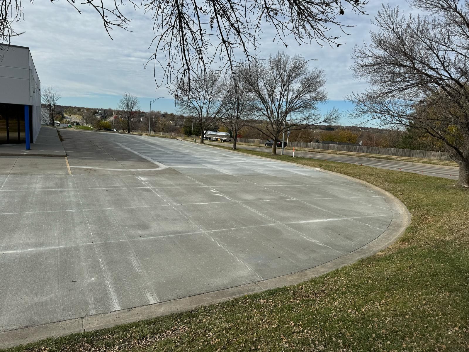 Empty concrete plaza beside a building, with grass, leafless trees, and a road in the background.