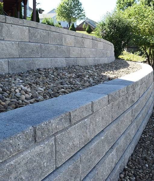 Curved gray stone retaining wall with gravel fill along a landscaped hillside.