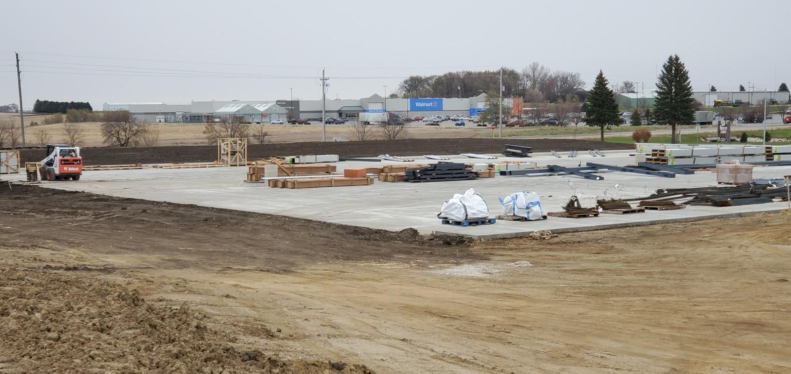 Construction site with excavated ground, concrete forms, and piles of materials under a cloudy sky