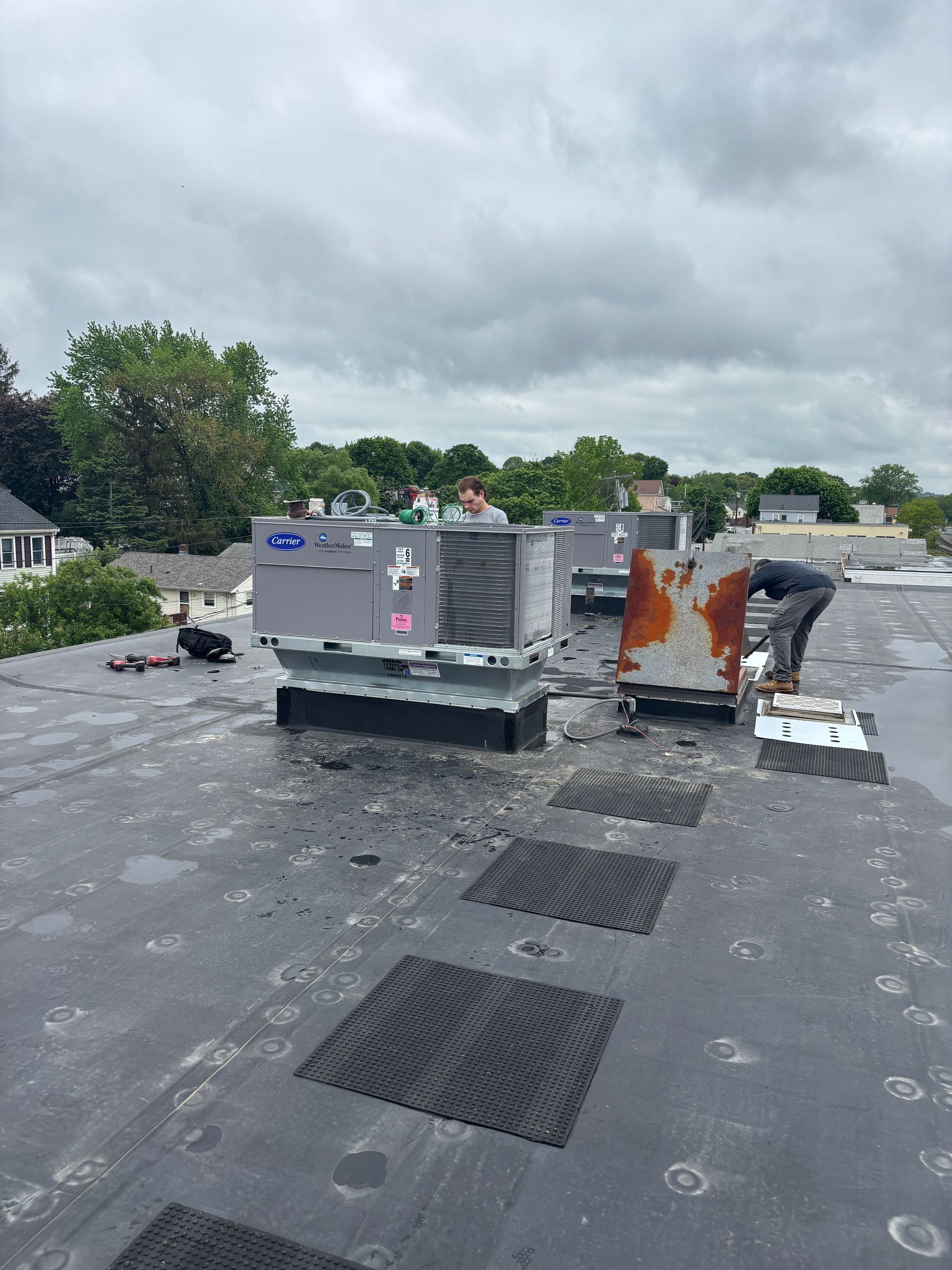 Workers service two HVAC units on a flat, dark gray commercial roof under a cloudy sky.