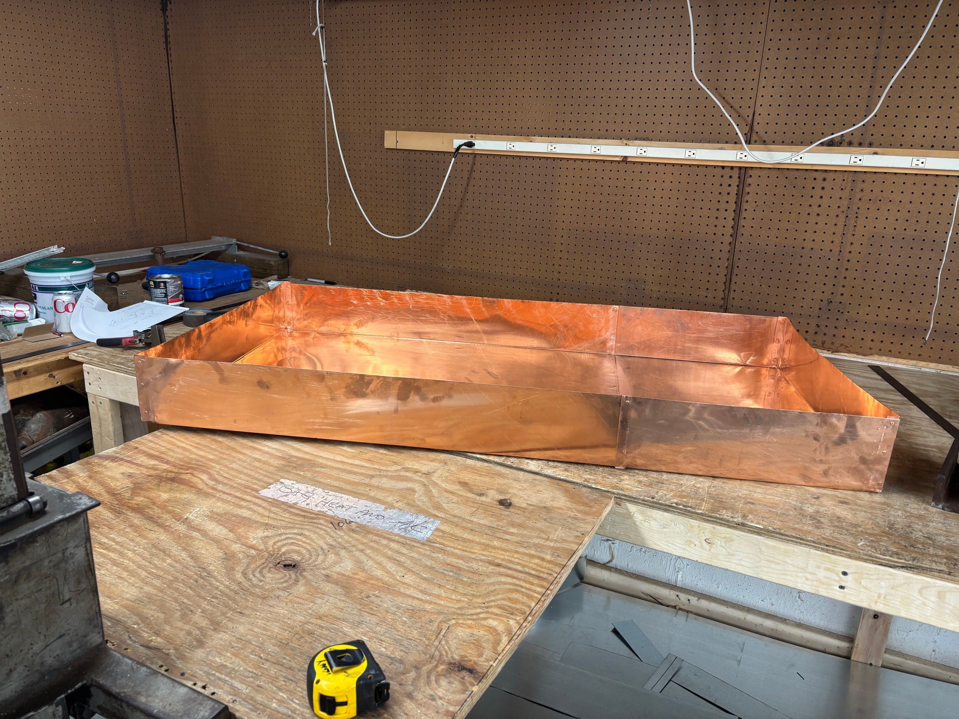 A rectangular copper pan sits on a workbench in a workshop, with a yellow tape measure and tools nearby.