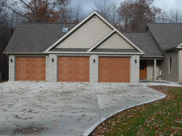 Three-car garage with tan siding and wooden doors, fronted by a wet concrete driveway and fallen leaves.