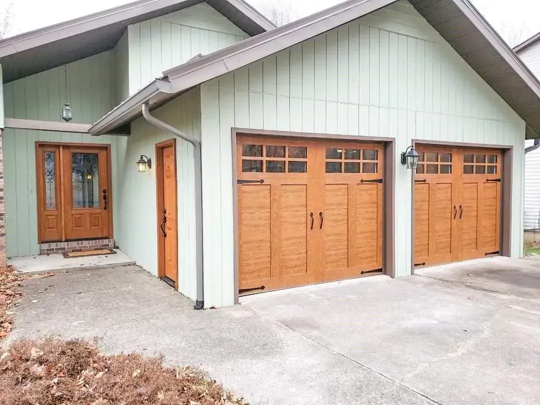 Exterior of a house with light green siding and three wooden garage doors beside a front entrance.