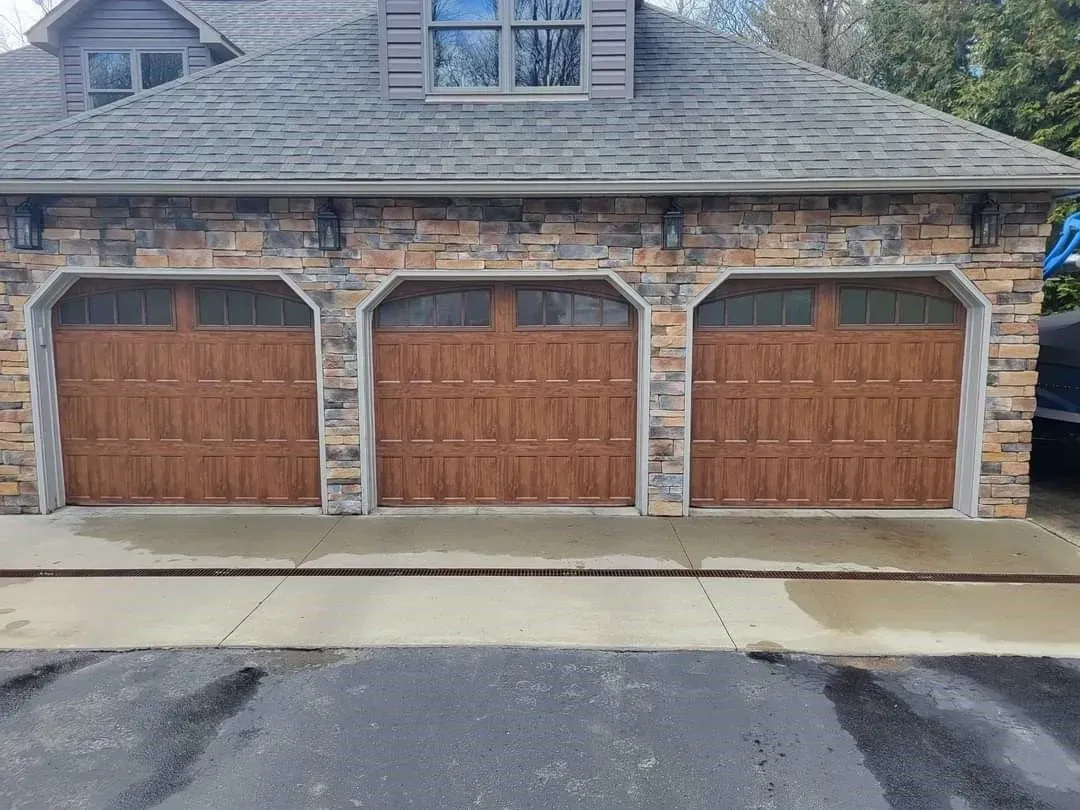 Three-car garage with brown wooden doors and stone facade beside a wet driveway