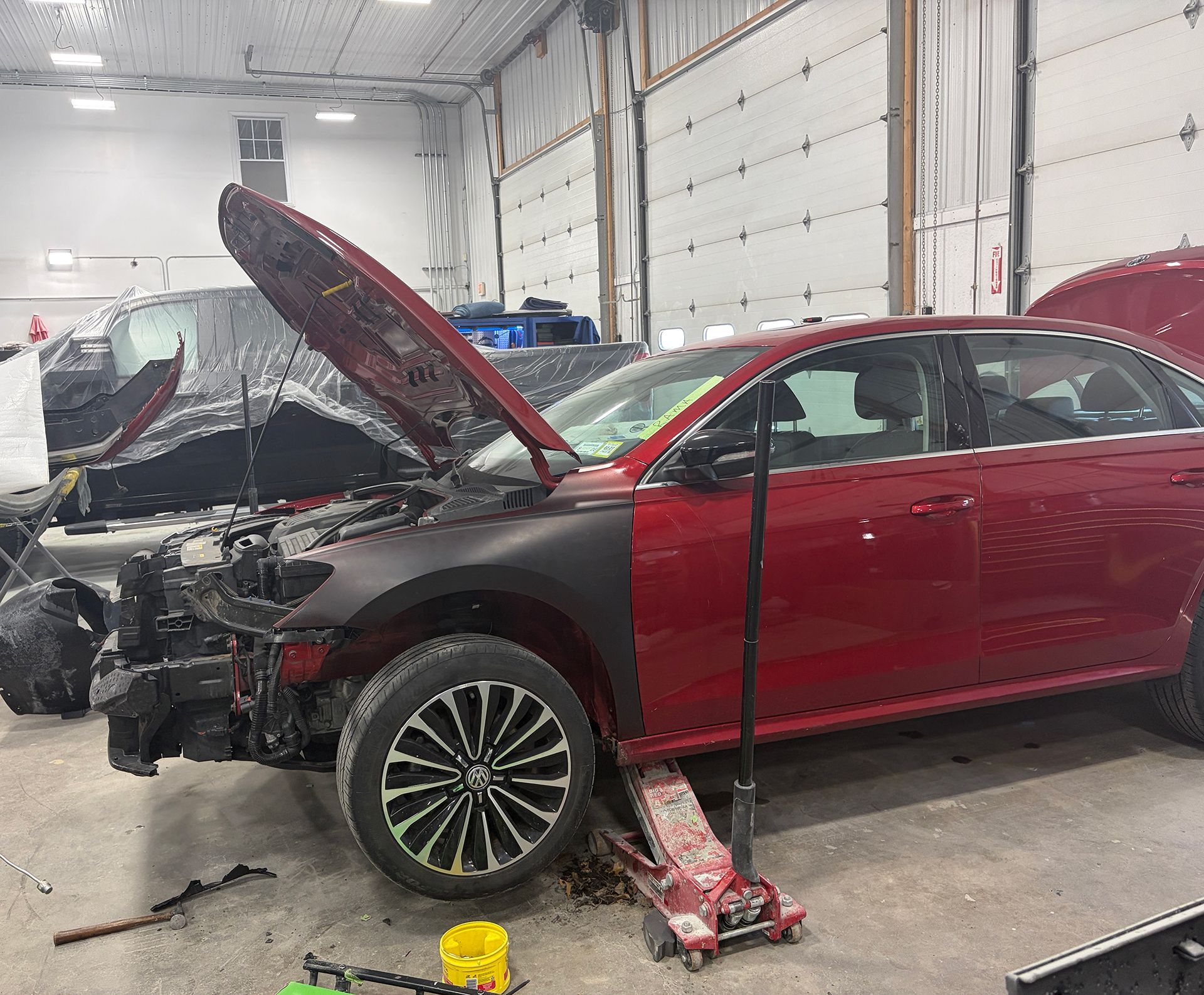 Damaged red sedan in a repair shop, hood open and front end severely wrecked
