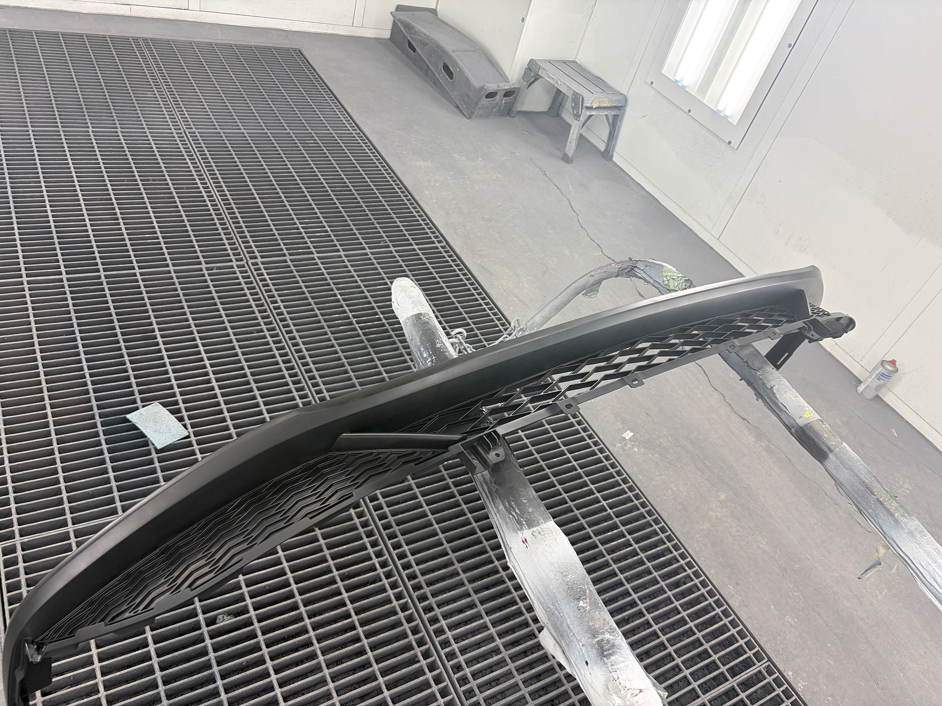 Black car bumper lying on a metal grate in a paint booth 