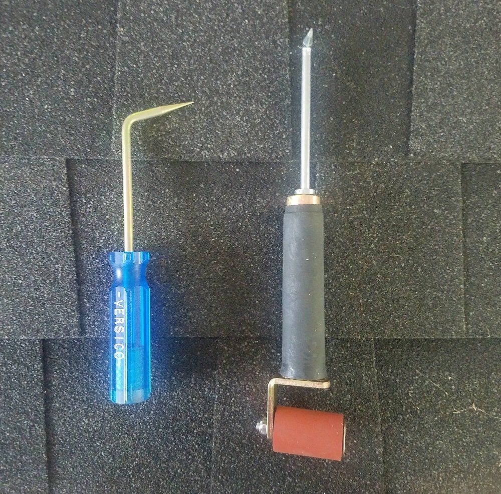 Two roofing tools: a hook and a pointed tool with a roller, on a shingle background.
