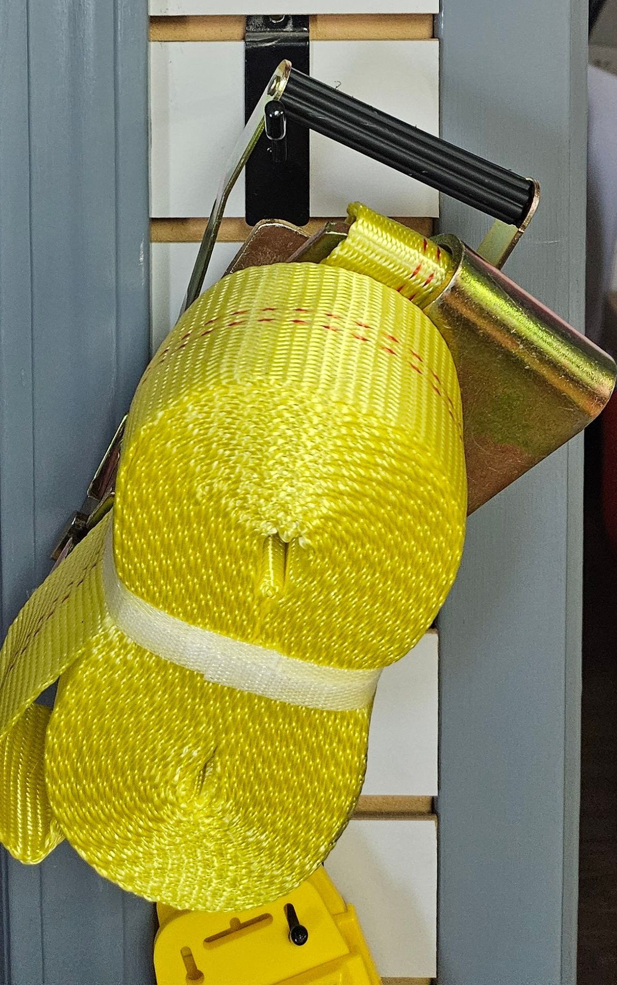Yellow cargo strap rolled up, hanging on a slat wall, with a metal handle.