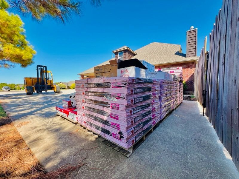 Pallets of pink insulation stacked next to a brick house under a blue sky, forklift in background.