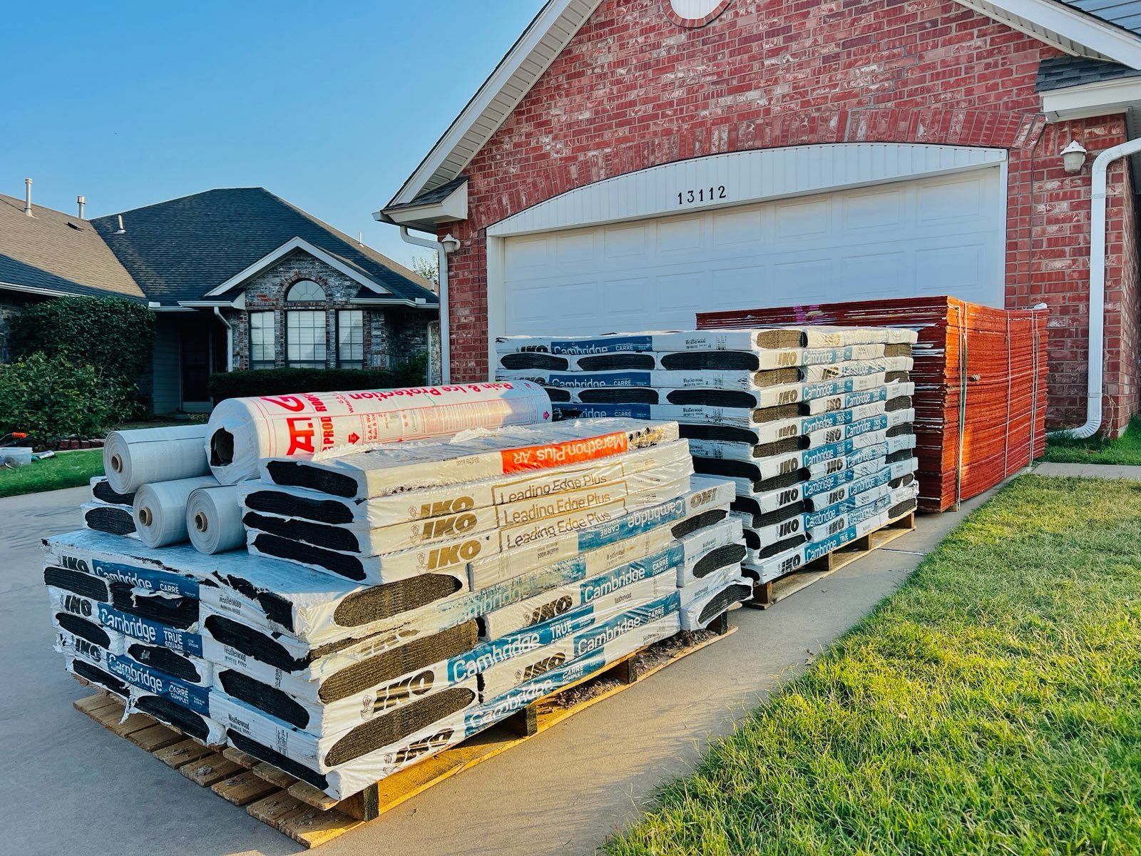 Pallets of roofing materials sit on a driveway in front of a brick house and garage.