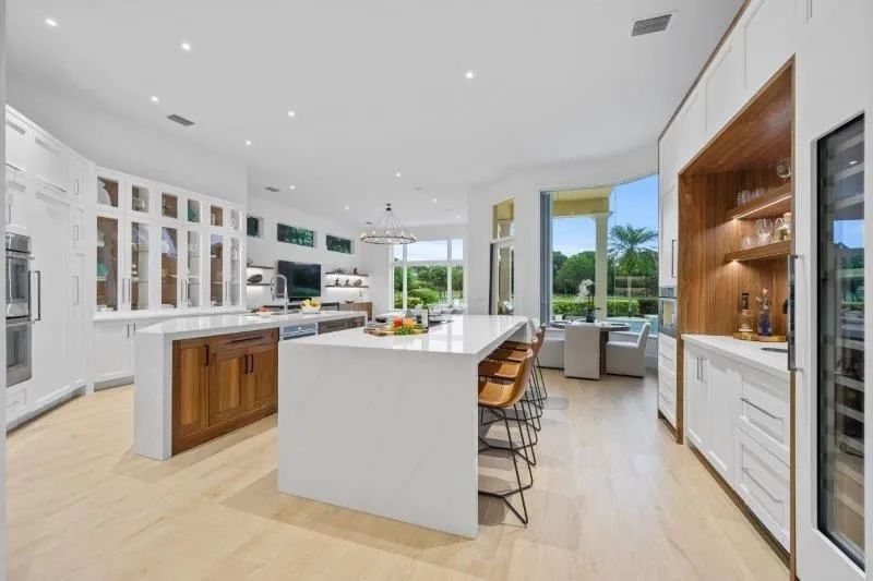 Modern white kitchen with island, wood accents, bar stools, and large windows overlooking greenery