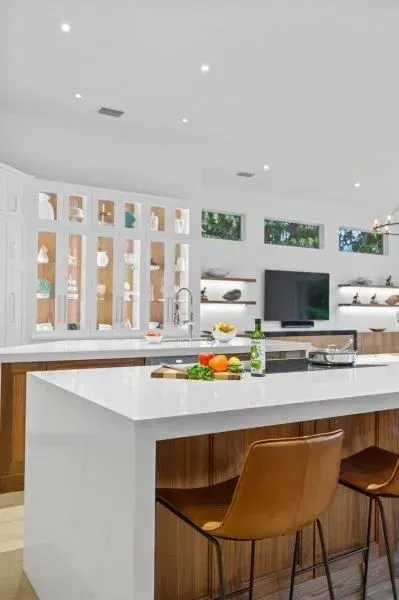 Bright modern kitchen with white island, wooden stools, and open view to a living area.