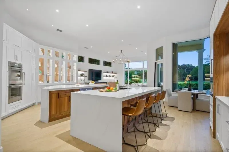 Modern white kitchen with large island, wood cabinets, pendant lights, and views of greenery outside