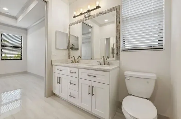 Bright white bathroom with vanity, large mirror, and toilet near a window.
