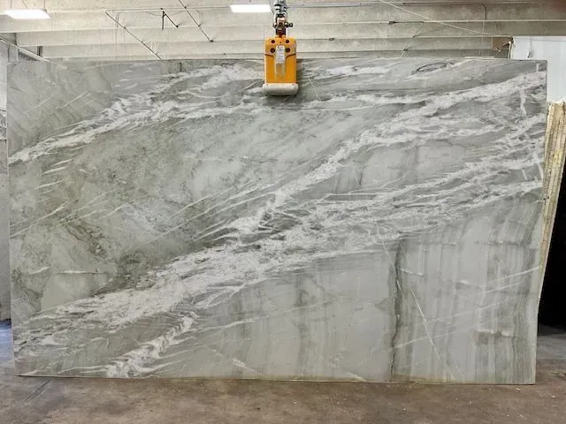 Large gray-and-white marble slab suspended by a yellow clamp in a warehouse