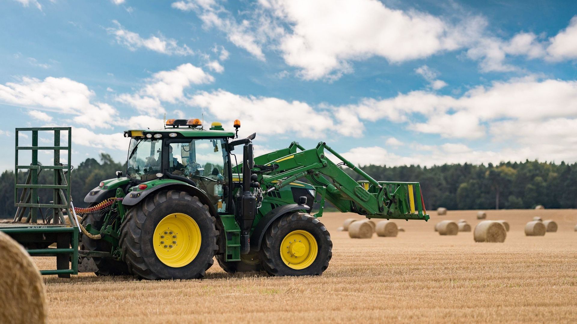 A green tractor is driving through a field of hay bales.