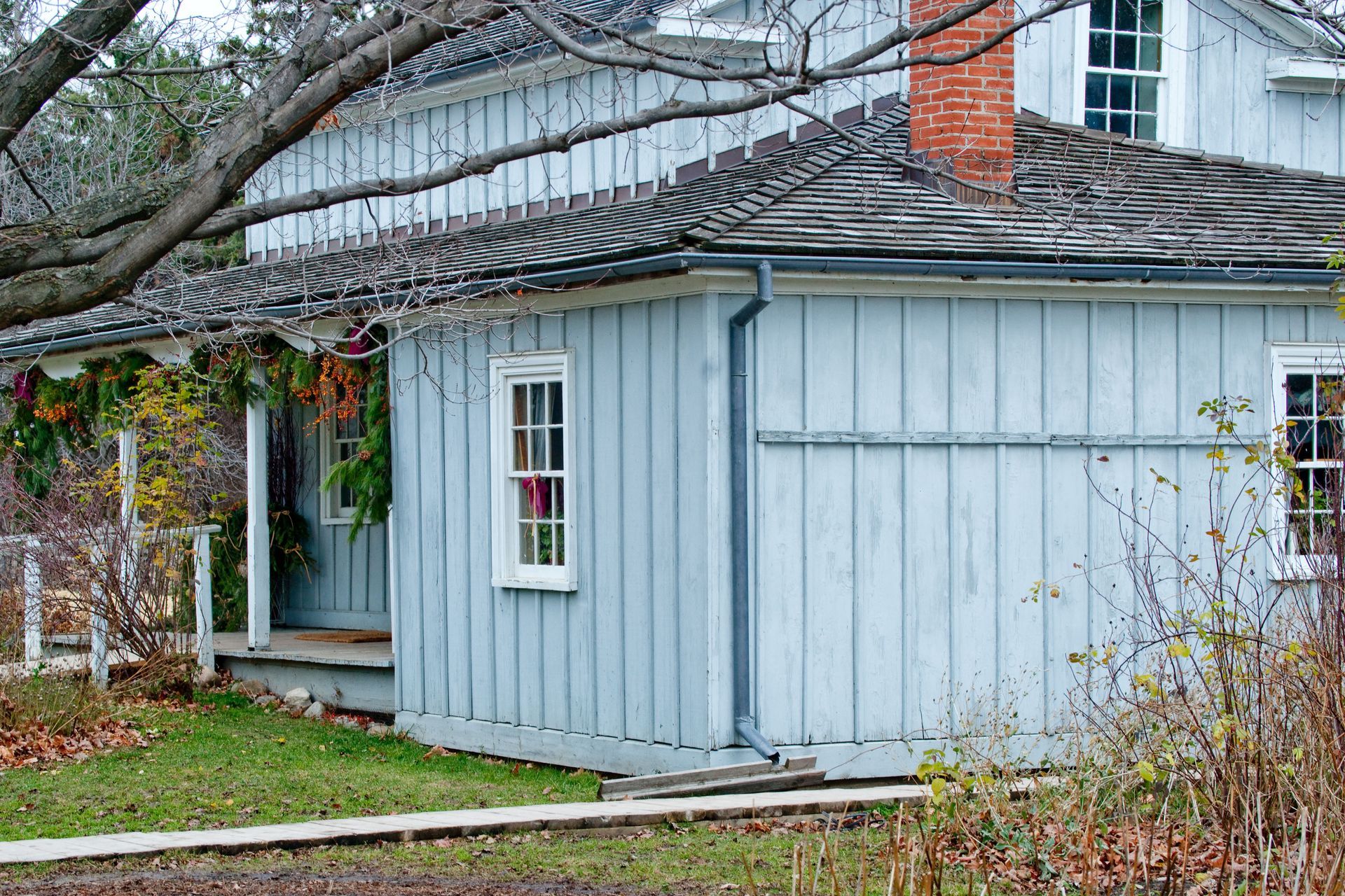 A rustic, light blue wooden house with a covered porch, a weathered shingle roof, and a red brick chimney.