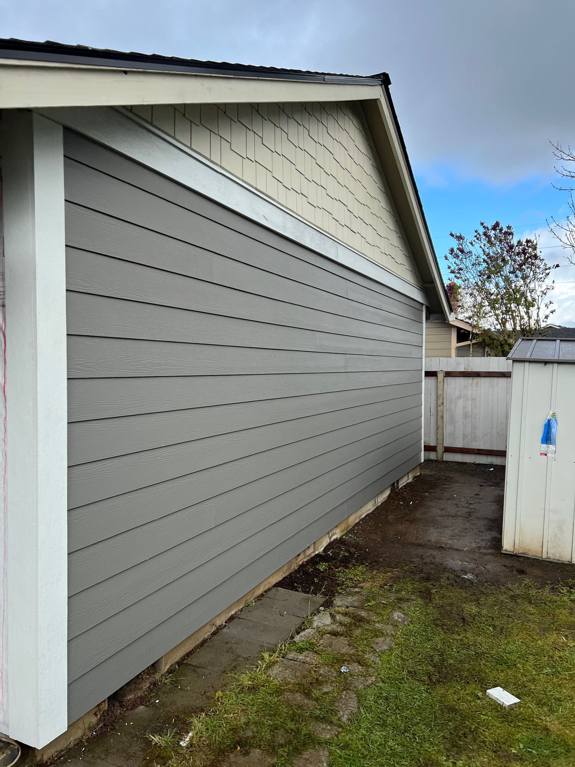 A side view of a house wall featuring new grey horizontal siding installed below the original off-white gable area.