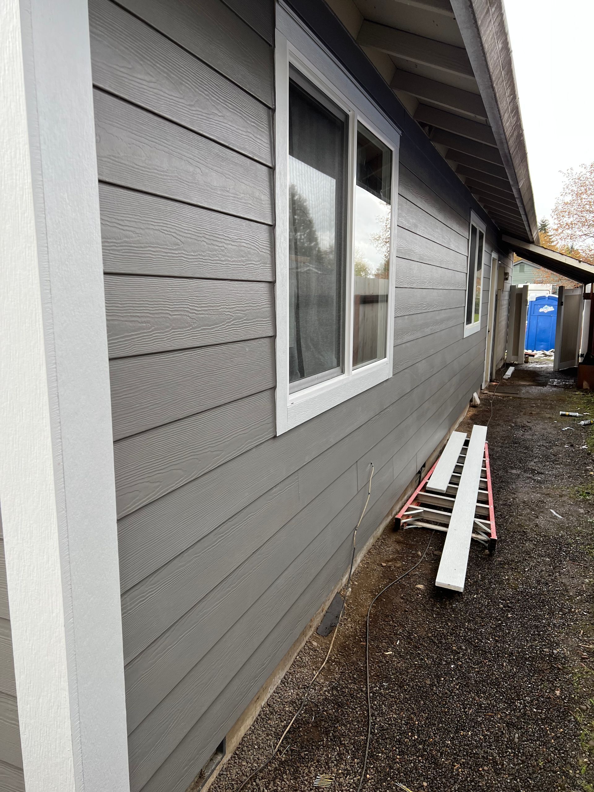 The side exterior of a house with grey horizontal siding, a white-trimmed window, and a ladder leaning against the wall.