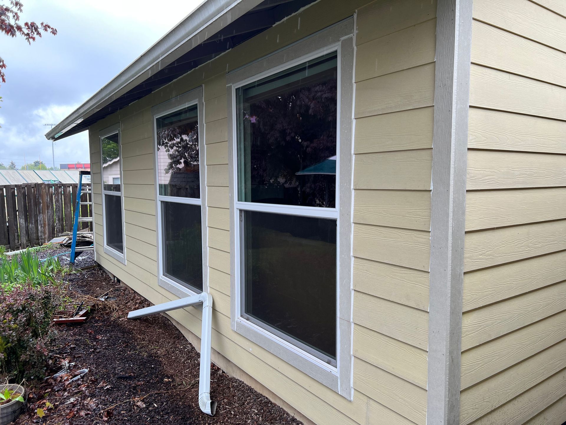 Side view of a light yellow house with three windows and a white downspout extension on a garden bed.
