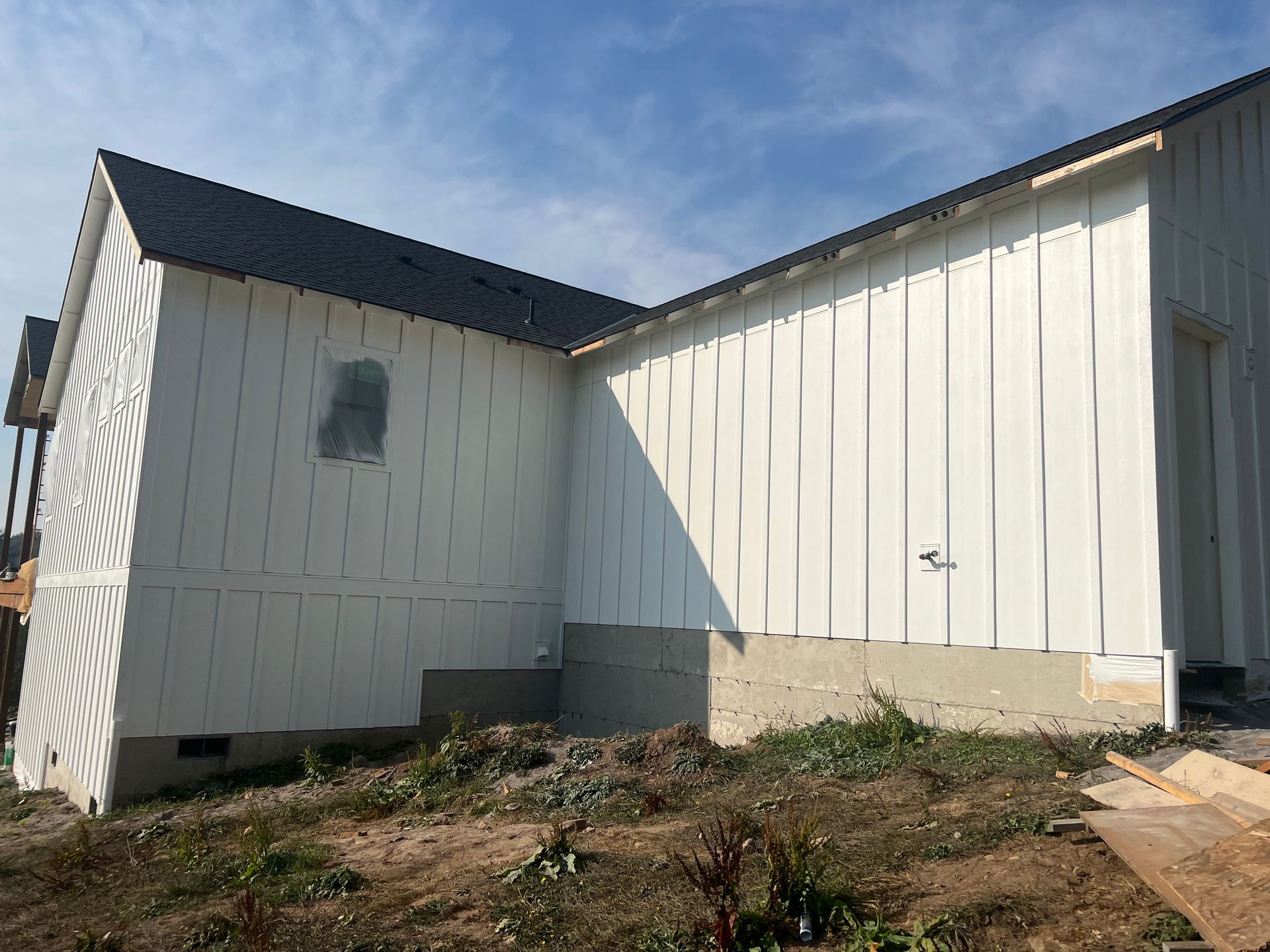 A residential building under construction with white vertical board-and-batten siding and a gray metal roof.
