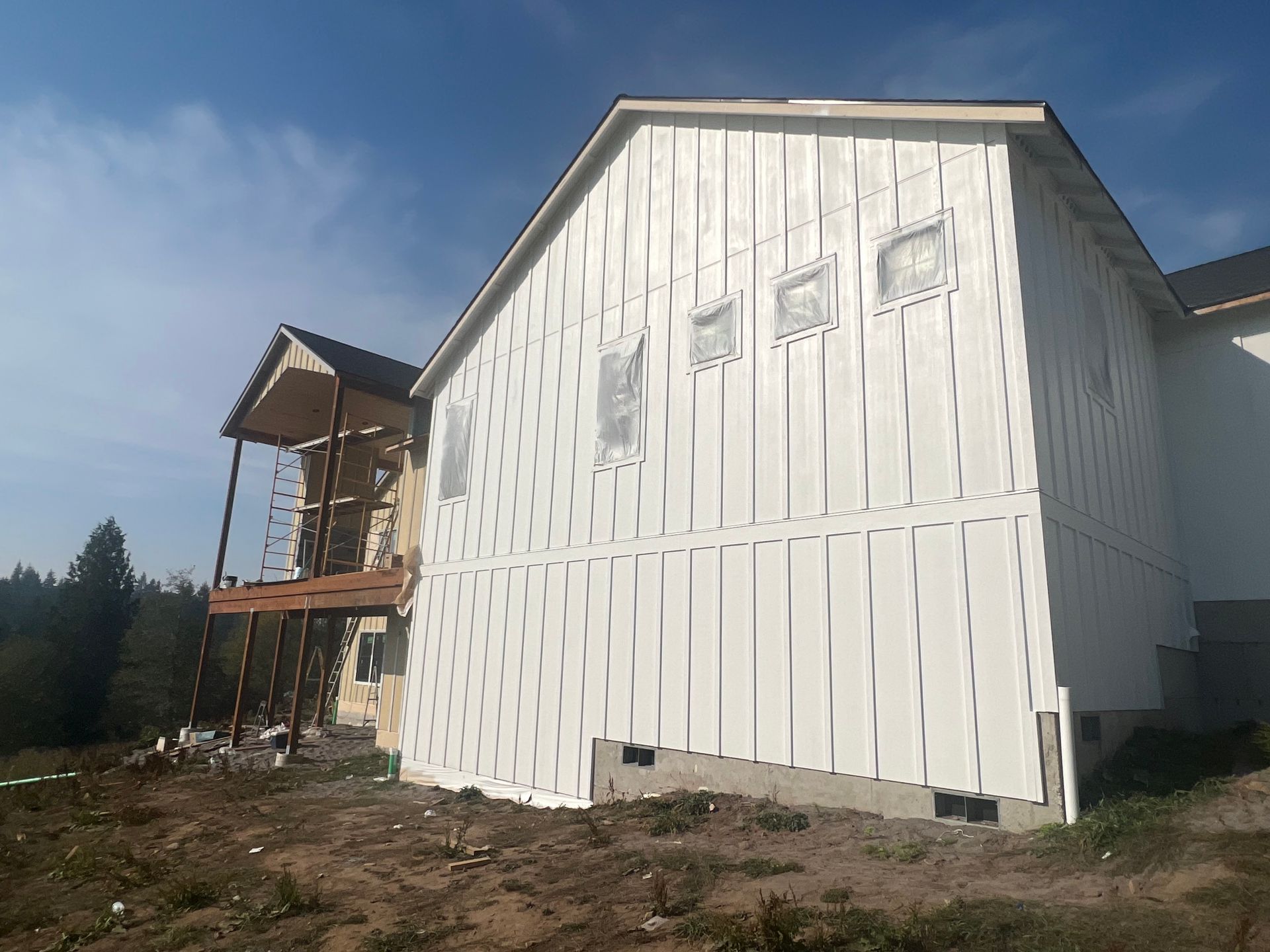 A side view of a house under construction with white vertical board-and-batten siding and a wooden deck on a hillside.