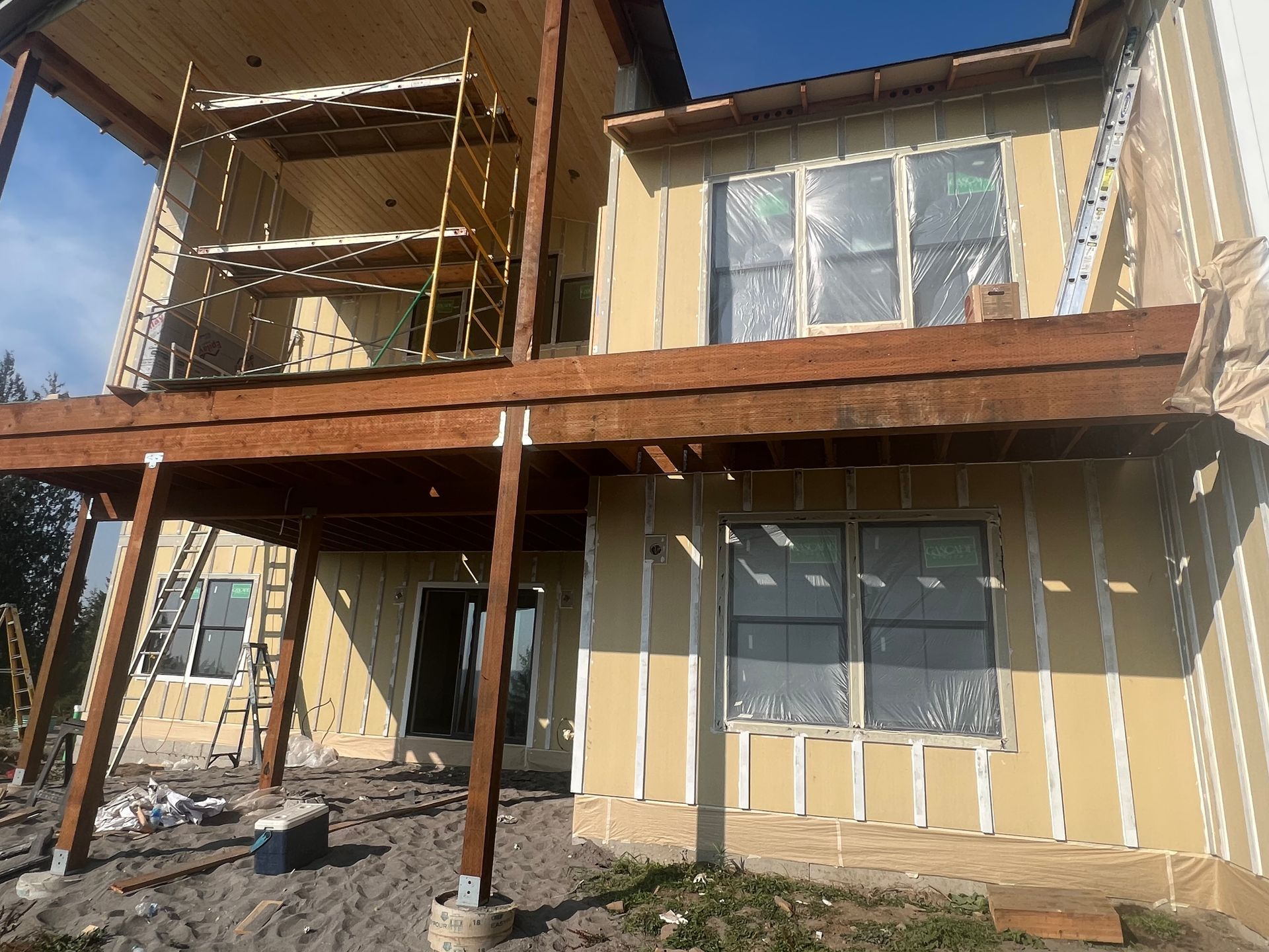 A multi-story house under construction with tan sheathing, installed windows, and a wooden deck frame and scaffolding.