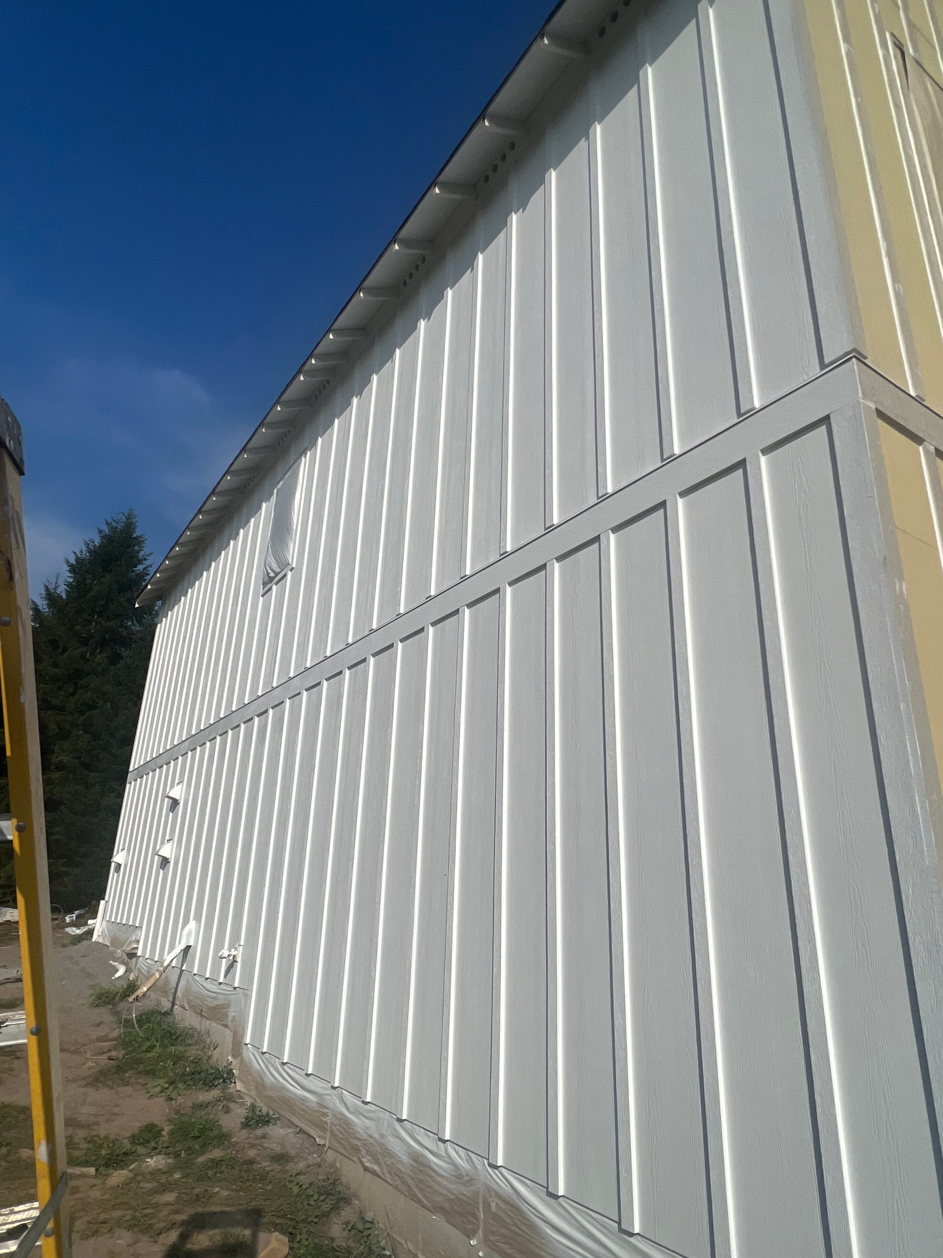 A side view of a building with white board-and-batten siding against a blue sky, with a yellow ladder in the foreground.