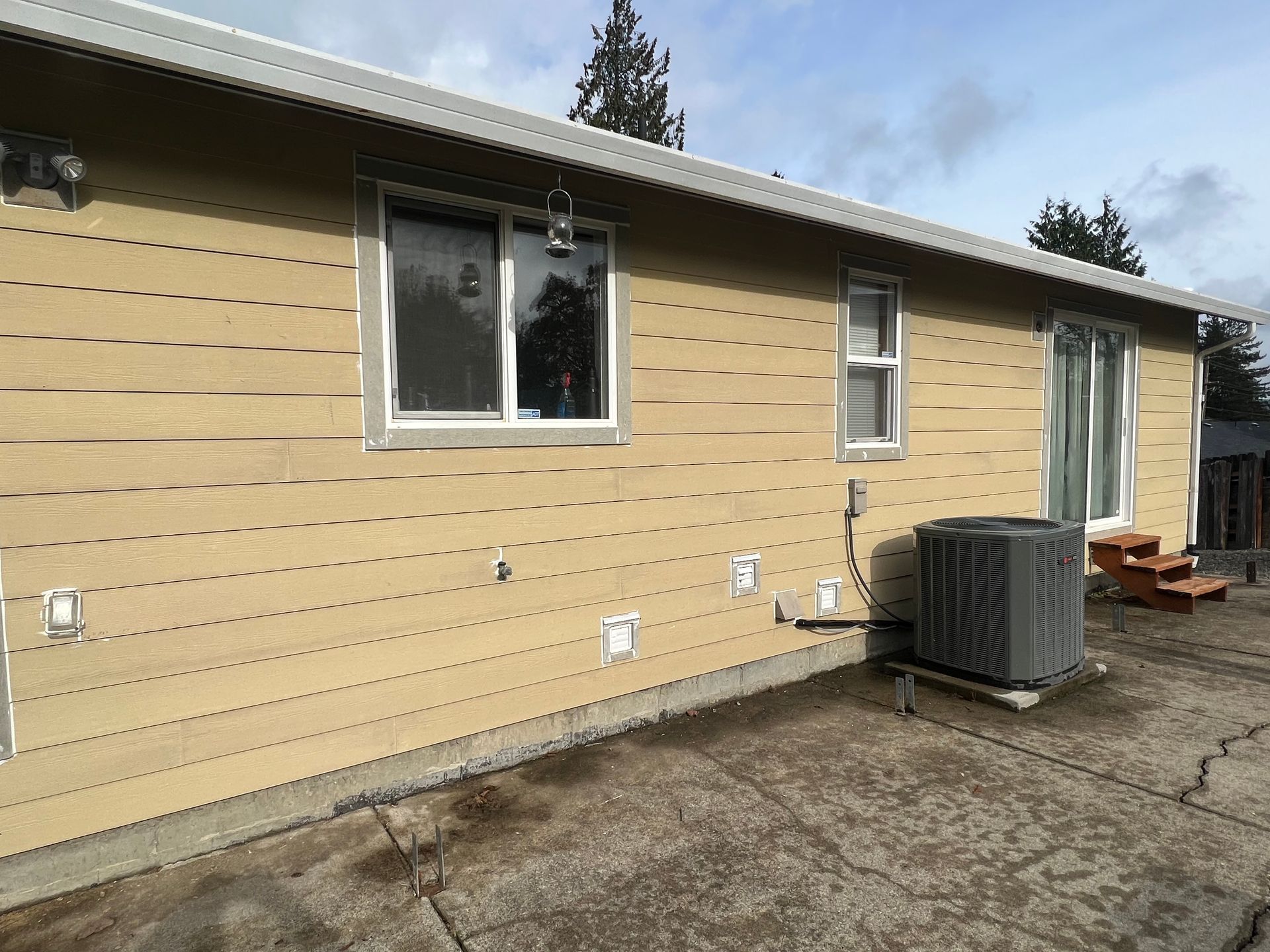 Exterior view of a house with tan horizontal siding, white-trimmed windows, a patio door, and an AC unit on a concrete pad.