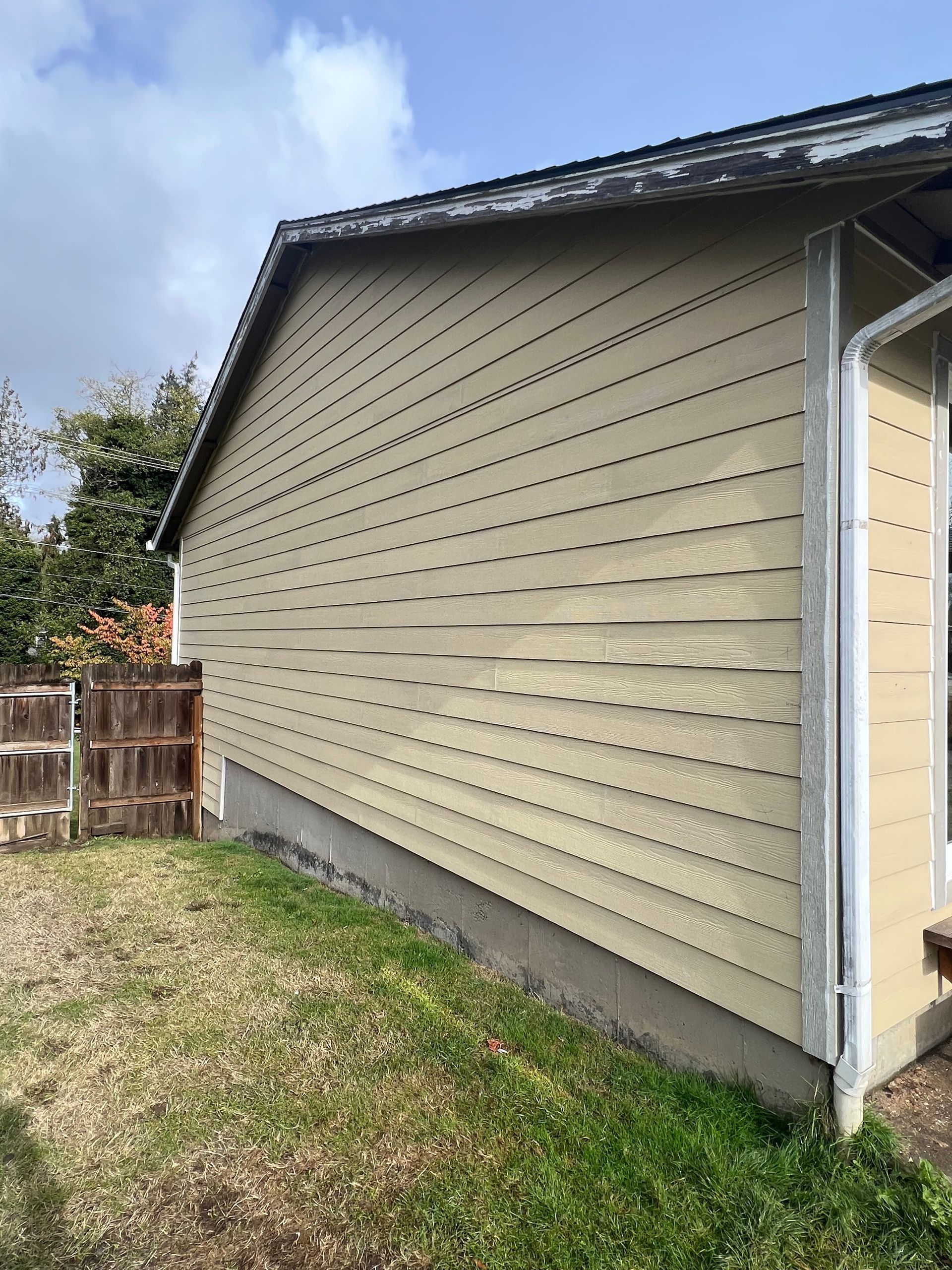 Side view of a house exterior with tan horizontal wood siding, showing noticeable signs of peeling paint and wear.
