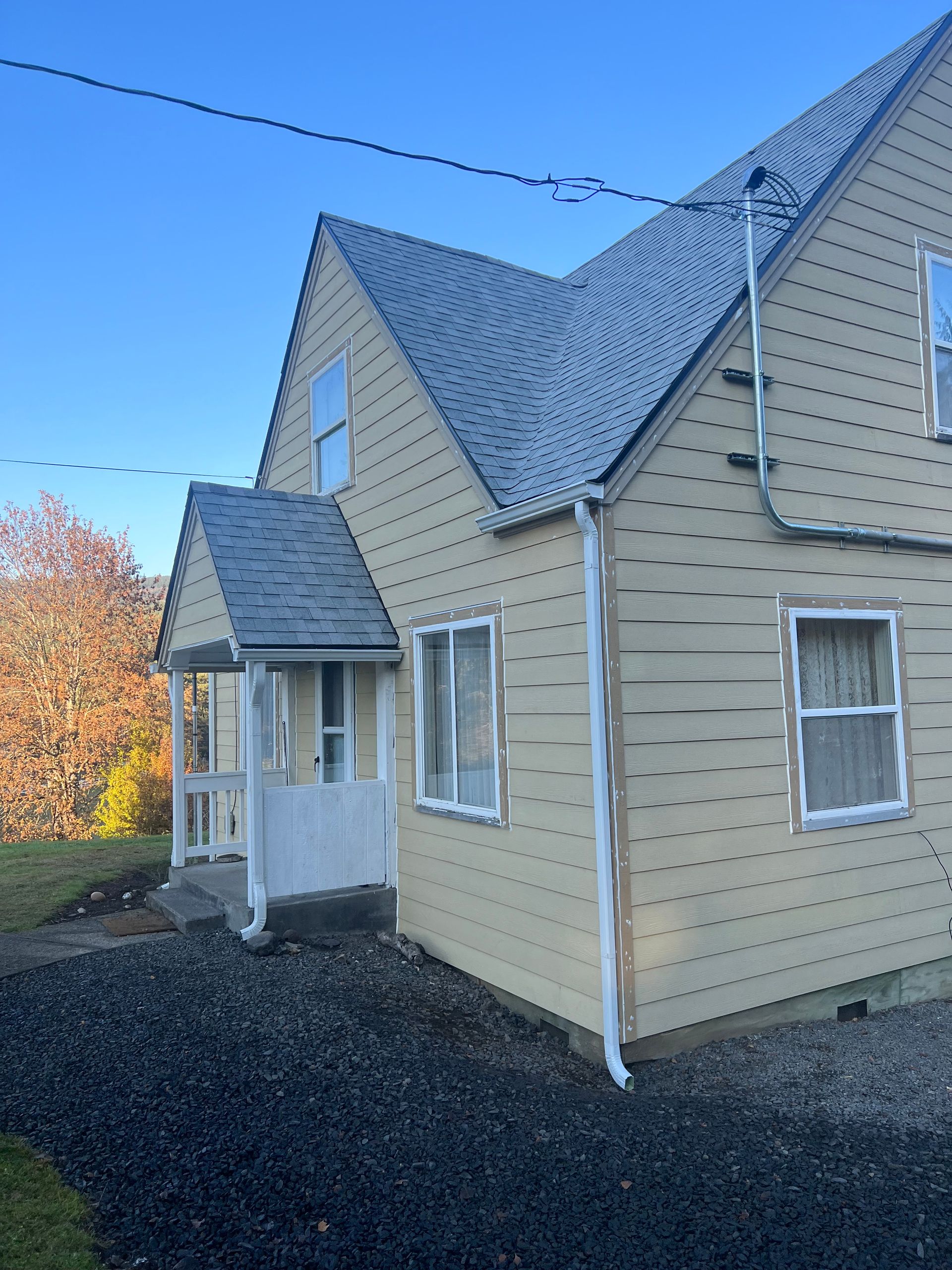 A yellow two-story house with a small front porch, dark shingled roof, and a gravel driveway on a sunny day.