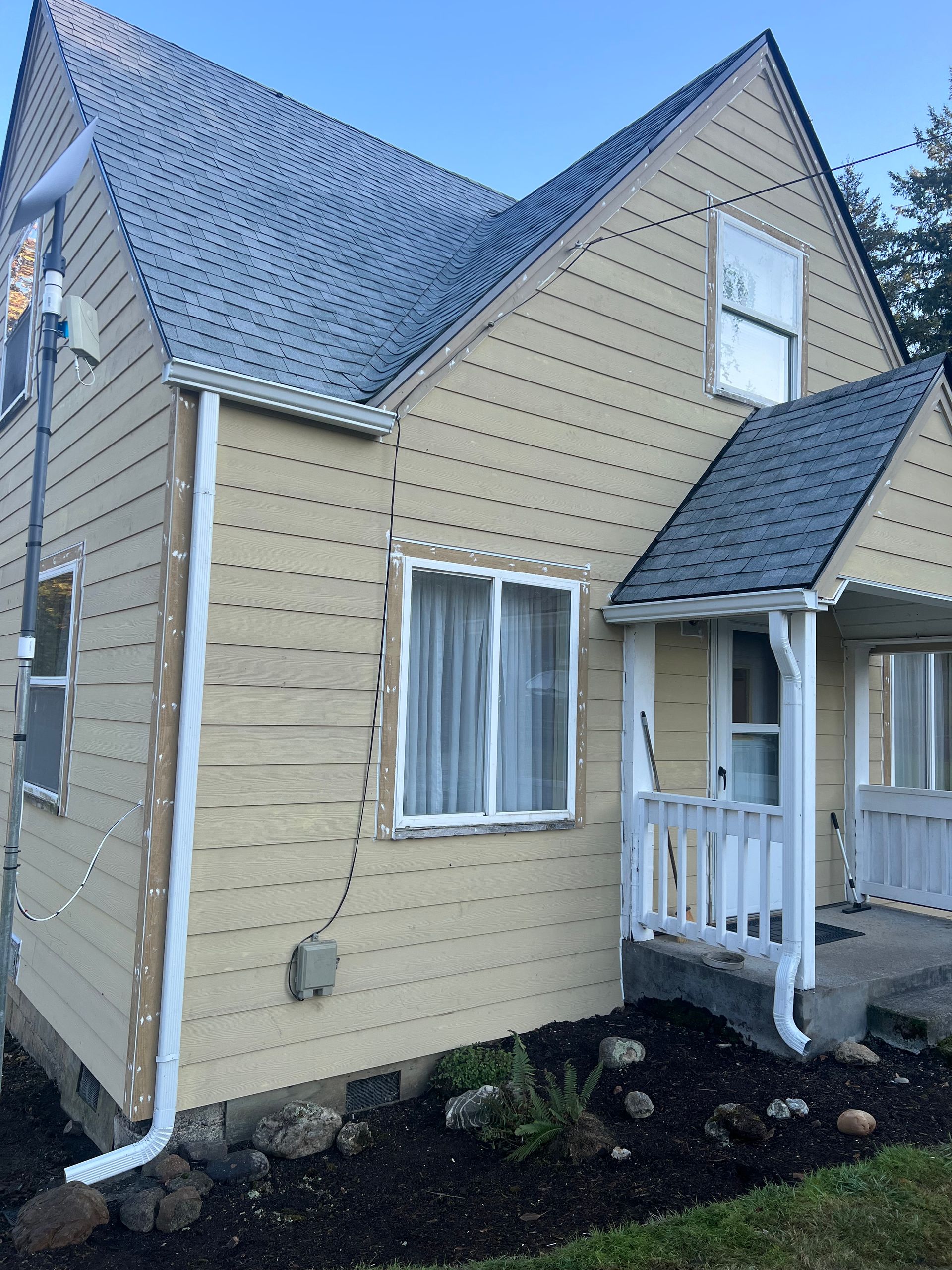 A tan, two-story house with a steep roof, white trim, a small front porch, and string lights hung along the exterior.